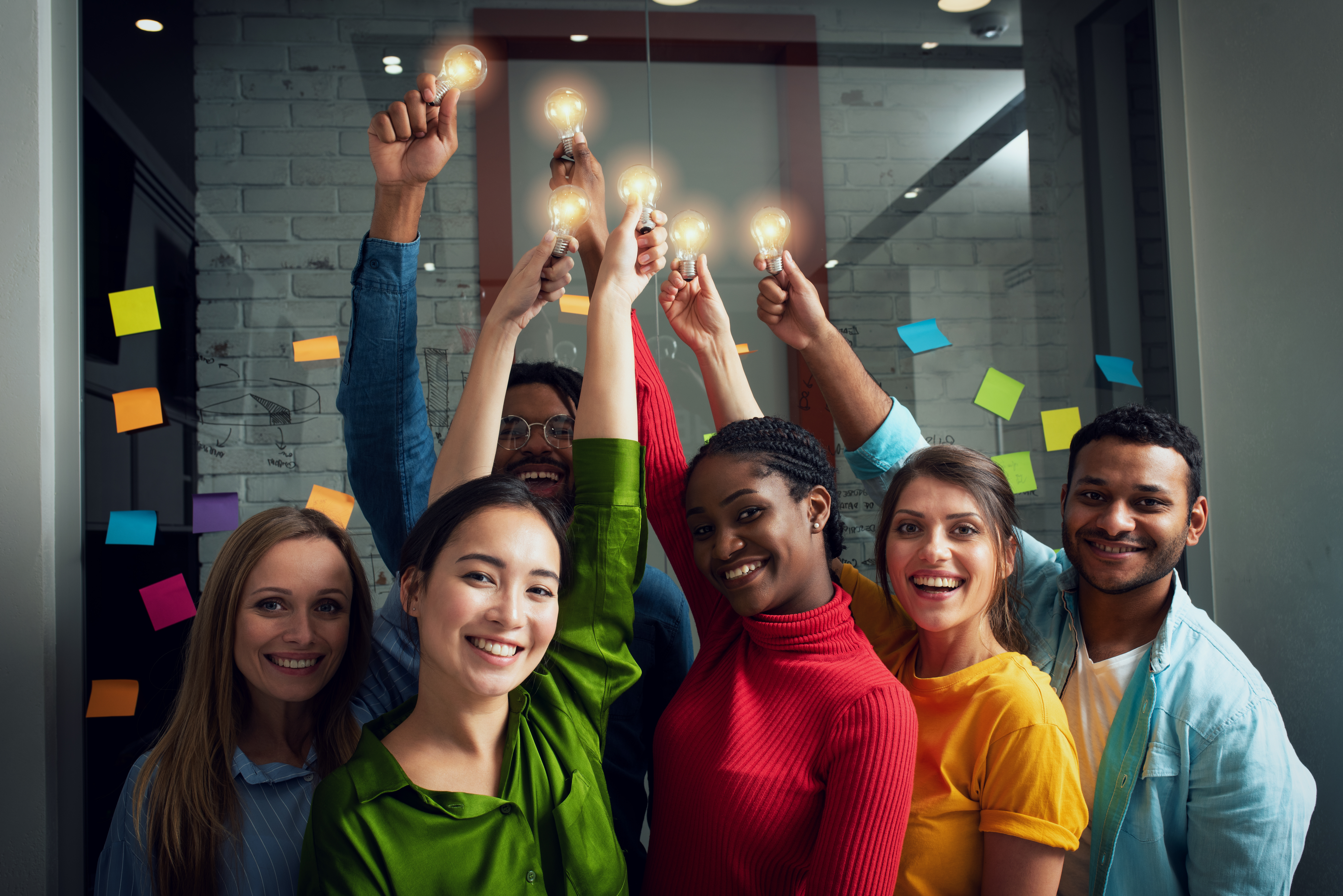 Grupo de jovens vestindo diferentes camisas coloridas segurando lâmpadas acesas e rodeados de post-its