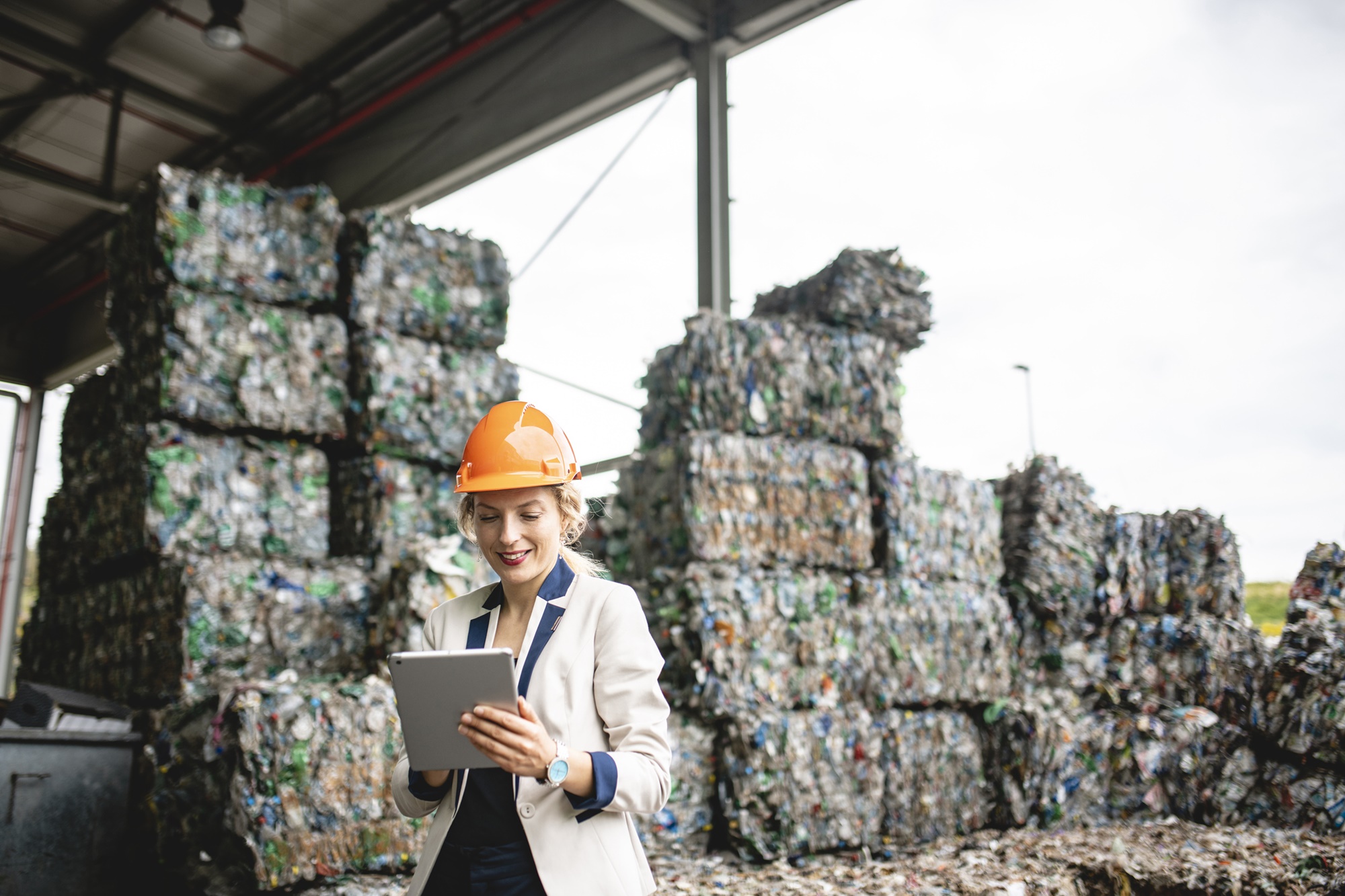 Mulher segurando tablet e vestindo capacete de proteção em frente a fardos de lixo reciclável.