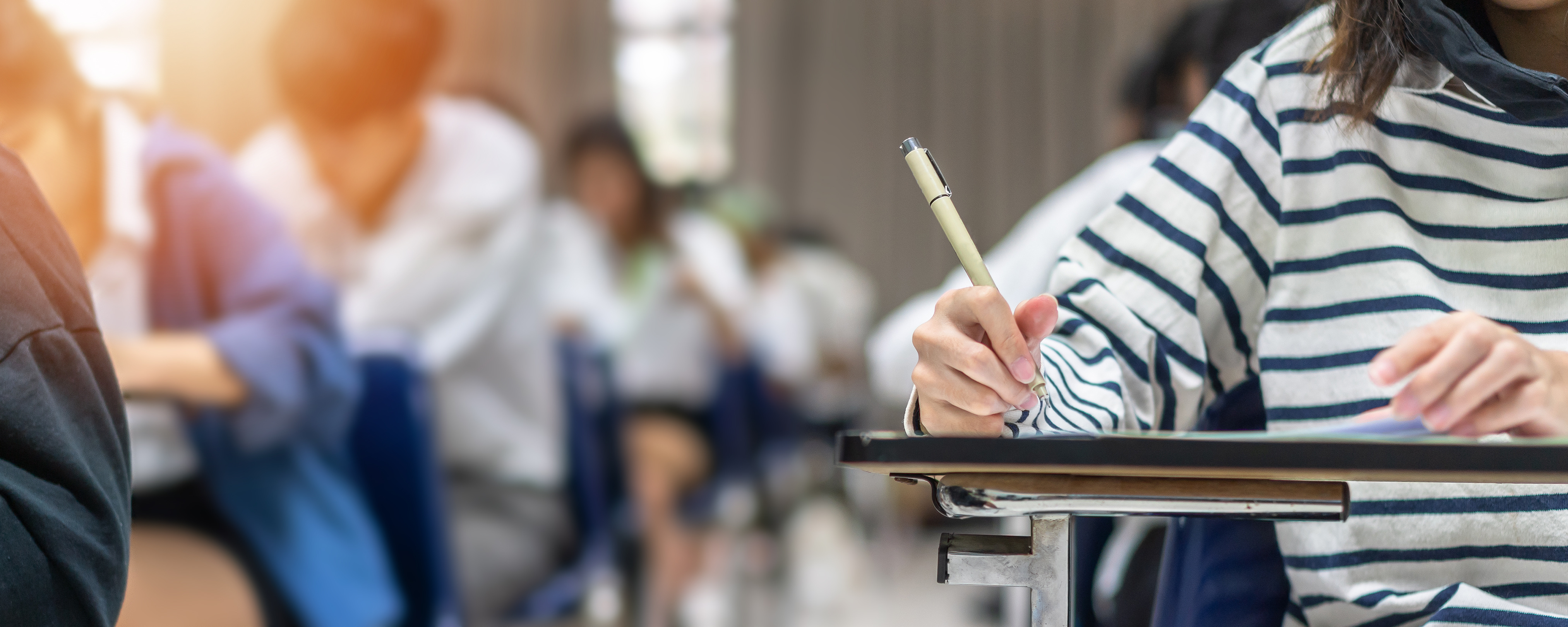 Imagem desfocada de uma sala de aula com algumas pessoas sentadas escrevendo.