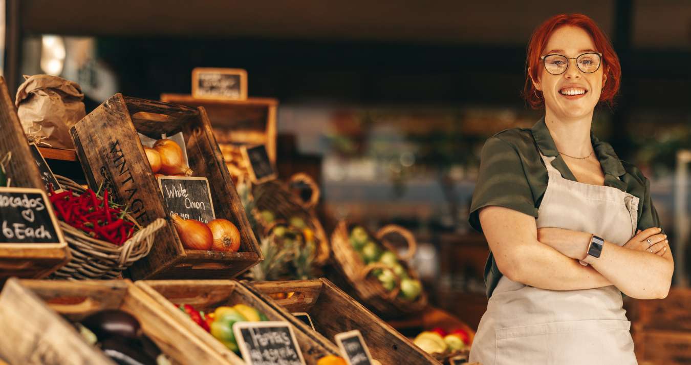 Mulher ruiva sorrindo com uma banca de frutas atrás