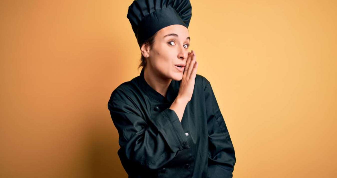 Jovem e chef de cozinha usando uniforme e chapéu, posando sobre um fundo amarelo, com a mão na boca contando um segredo ou boato, sussurrando uma conversa