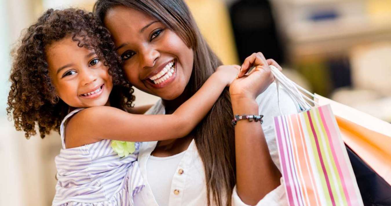 Foto de uma Mãe segurando uma sacola de presente e sua filha sorrindo e se abraçamdo