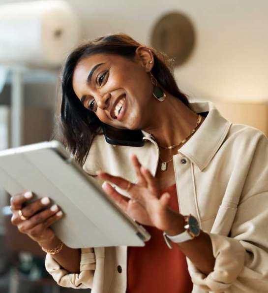Uma mulher empreendedora sorridente conversa pelo telefone enquanto segura um tablet. Ela está bem vestida com uma blusa clara, usando joias elegantes, e parece estar em um espaço de trabalho interno com caixas empilhadas ao fundo, sugerindo um ambiente de negócios ou logística.