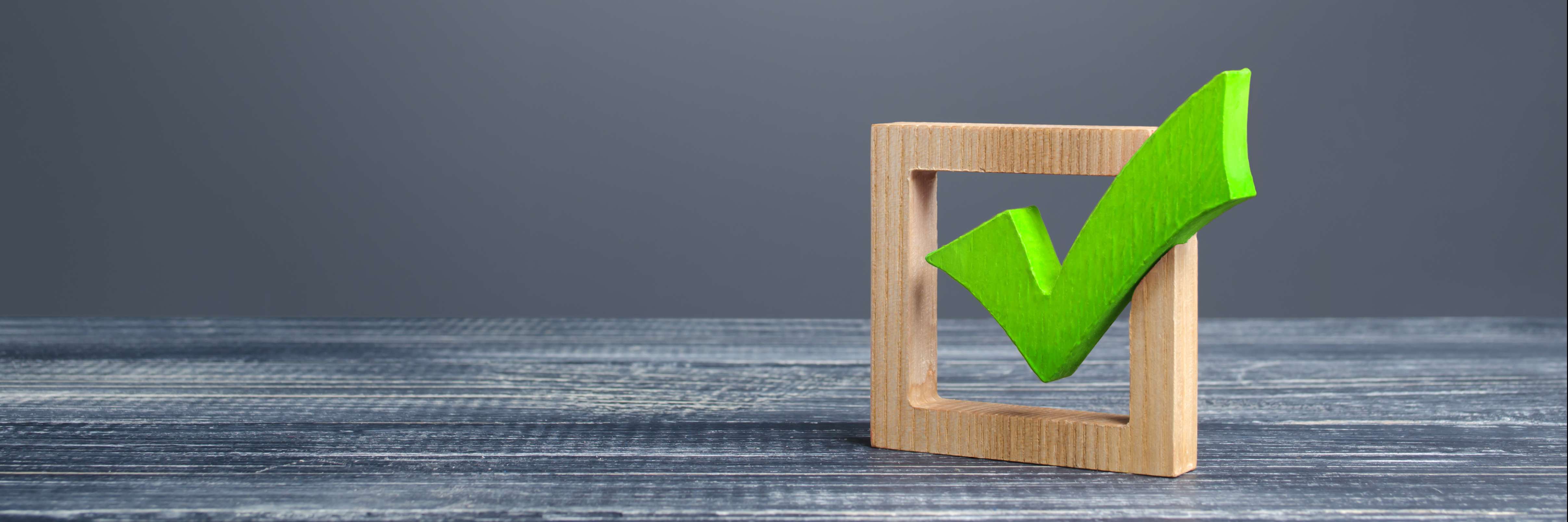 Quadrado de madeira, como se fosse uma caixa de seleção, e um "check" em verde dentro do quadrado, ambos apoiados em uma superfície. 