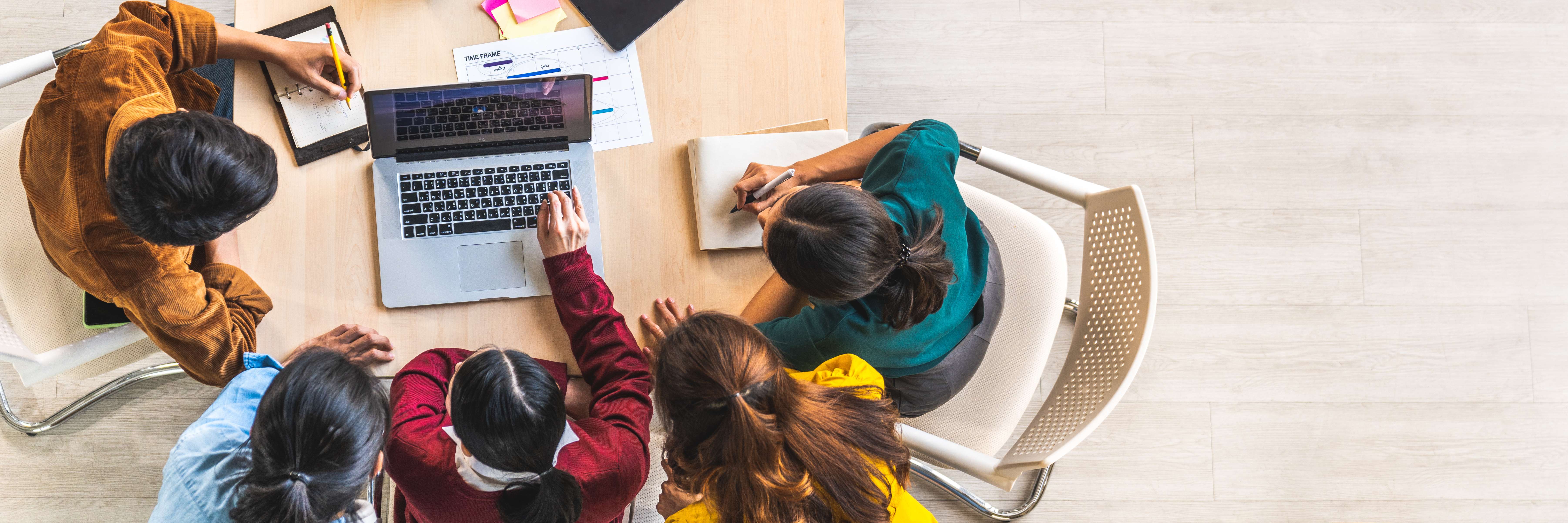 Cinco pessoas em uma mesa com notebook, tablet e papéis, vistos de cima.