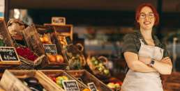 Mulher ruiva sorrindo com uma banca de frutas atrás